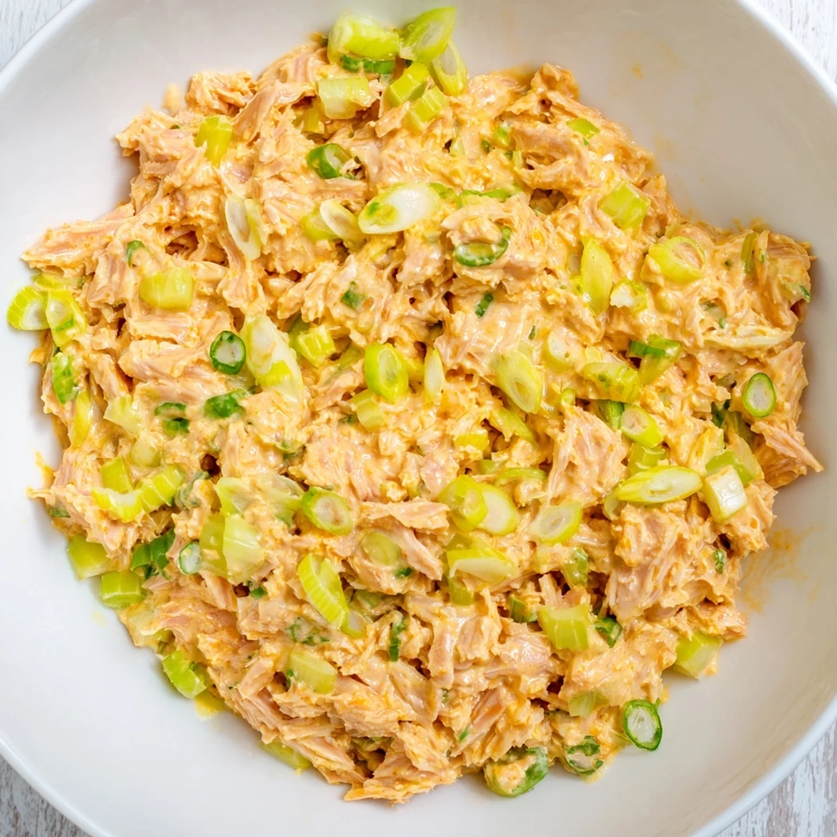 Canned tuna chunks coated in creamy spicy mayo, dotted with sliced scallions and celery in a white bowl.